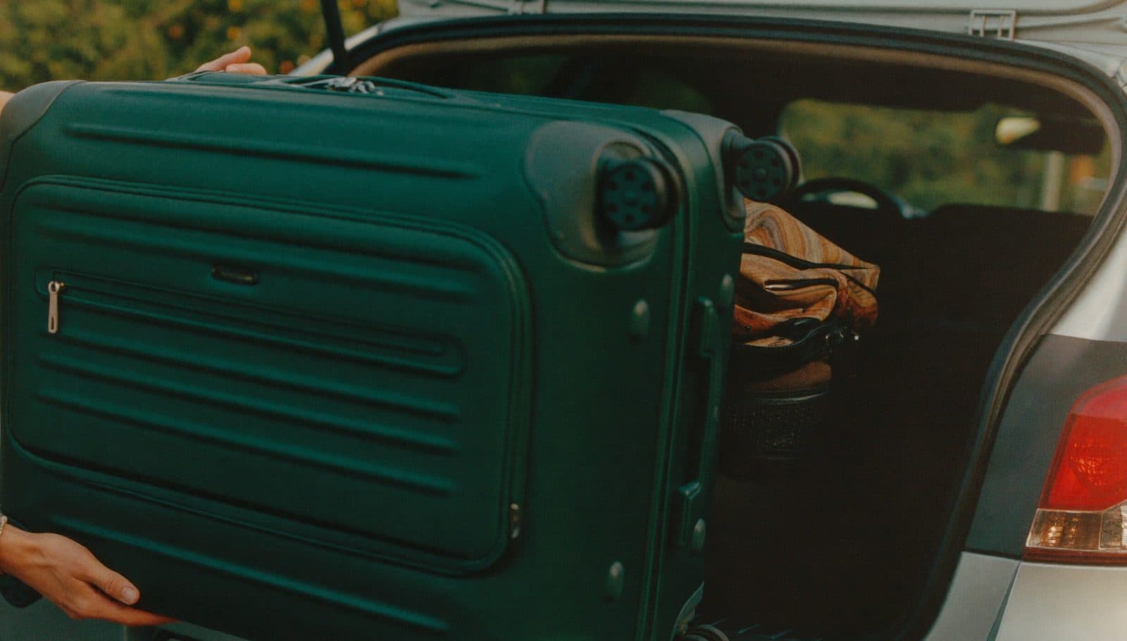 Hands lifting a suitcase into the open boot of a car at the kerbside in warm afternoon light